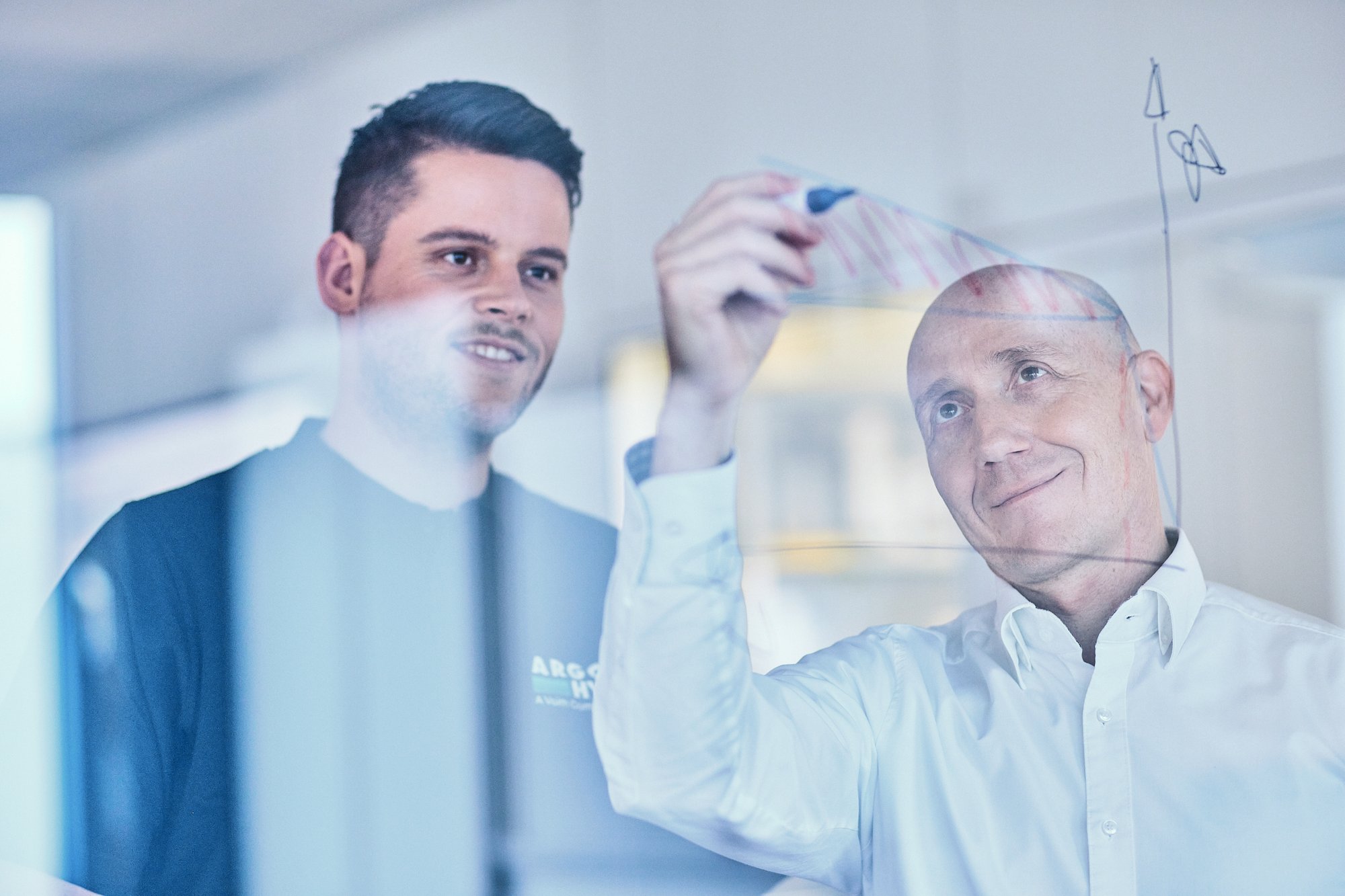 Zwei Mitarbeitende skizzieren und besprechen technische Konzepte auf einer Glaswand. Zwei Mitarbeitende skizzieren und besprechen technische Konzepte auf einer Glaswand.