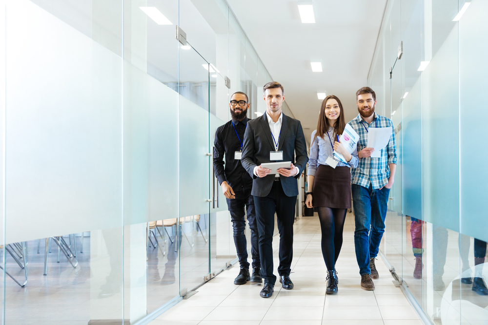 Four employees walking through a modern office corridor carrying documents and a tablet.