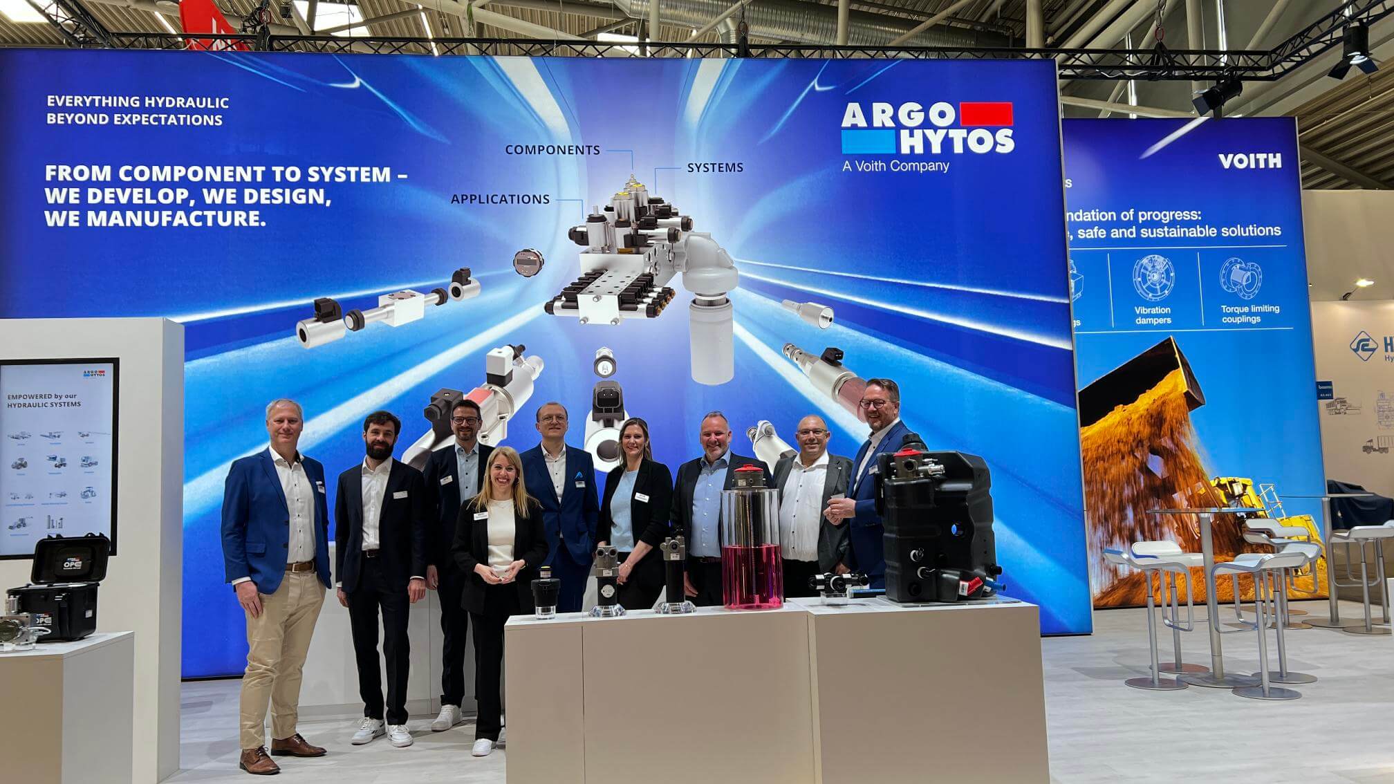 Gruppenfoto vor einem ARGO-HYTOS Messestand mit ausgestellten Hydraulikkomponenten und großflächigen Produktgrafiken.