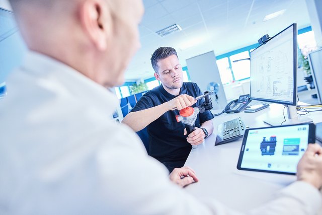 Two employees reviewing a technical component and CAD design on a monitor and tablet in an office workspace. Two employees reviewing a technical component and CAD design on a monitor and tablet in an office workspace.