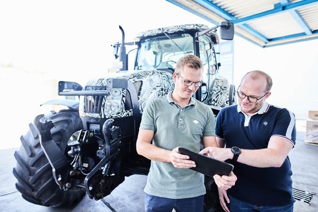 Zwei Mitarbeitende prüfen Dokumente auf einem Tablet vor einem Traktor in einer Werkstatt.